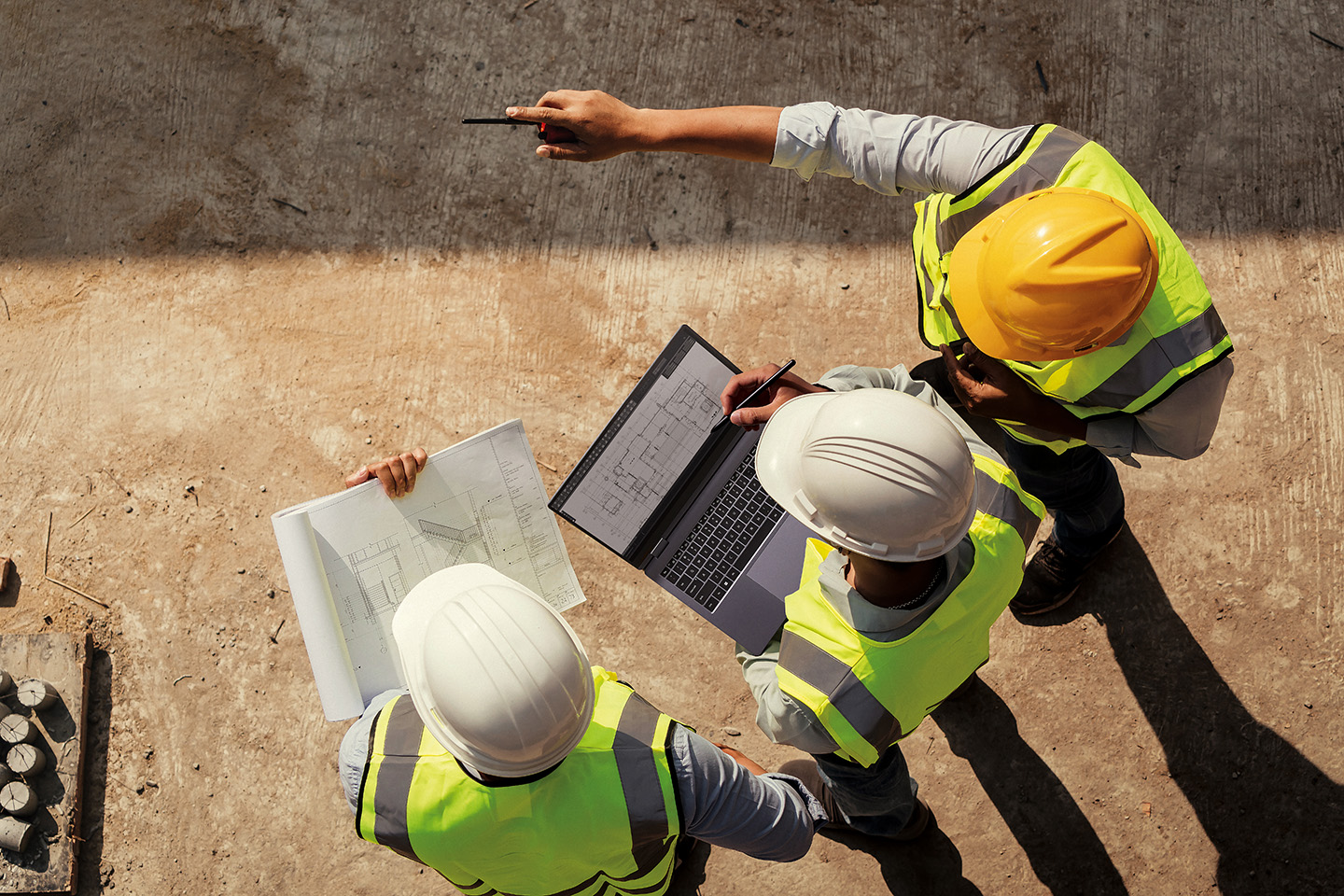 An overhead view of field workers in high-visibility vests reviewing construction drawings on a laptop and paper plans at a job site.