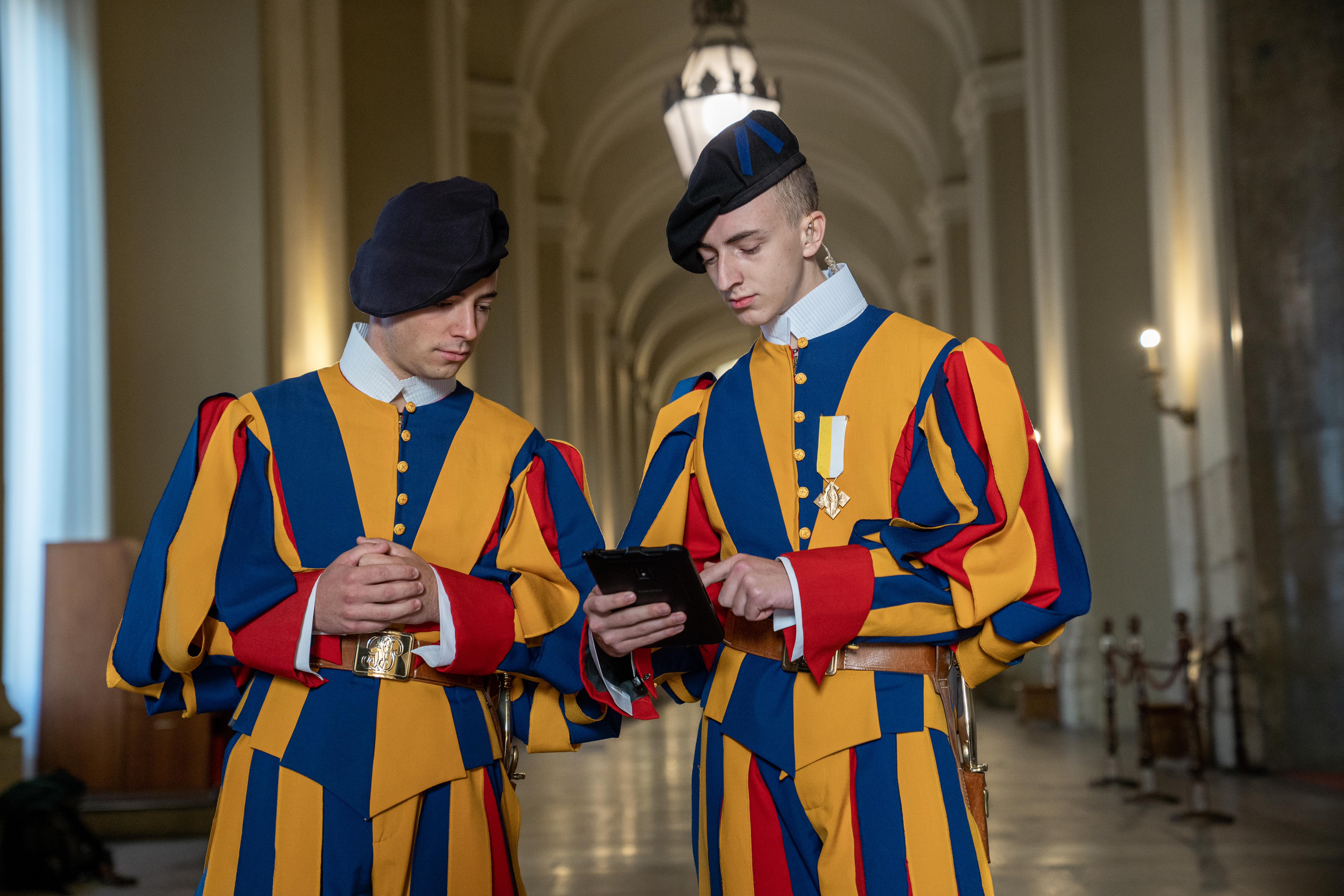 A pair of Vatican Swiss Guards standing in a vaulted hallway while looking at a Samsung rugged devices.
