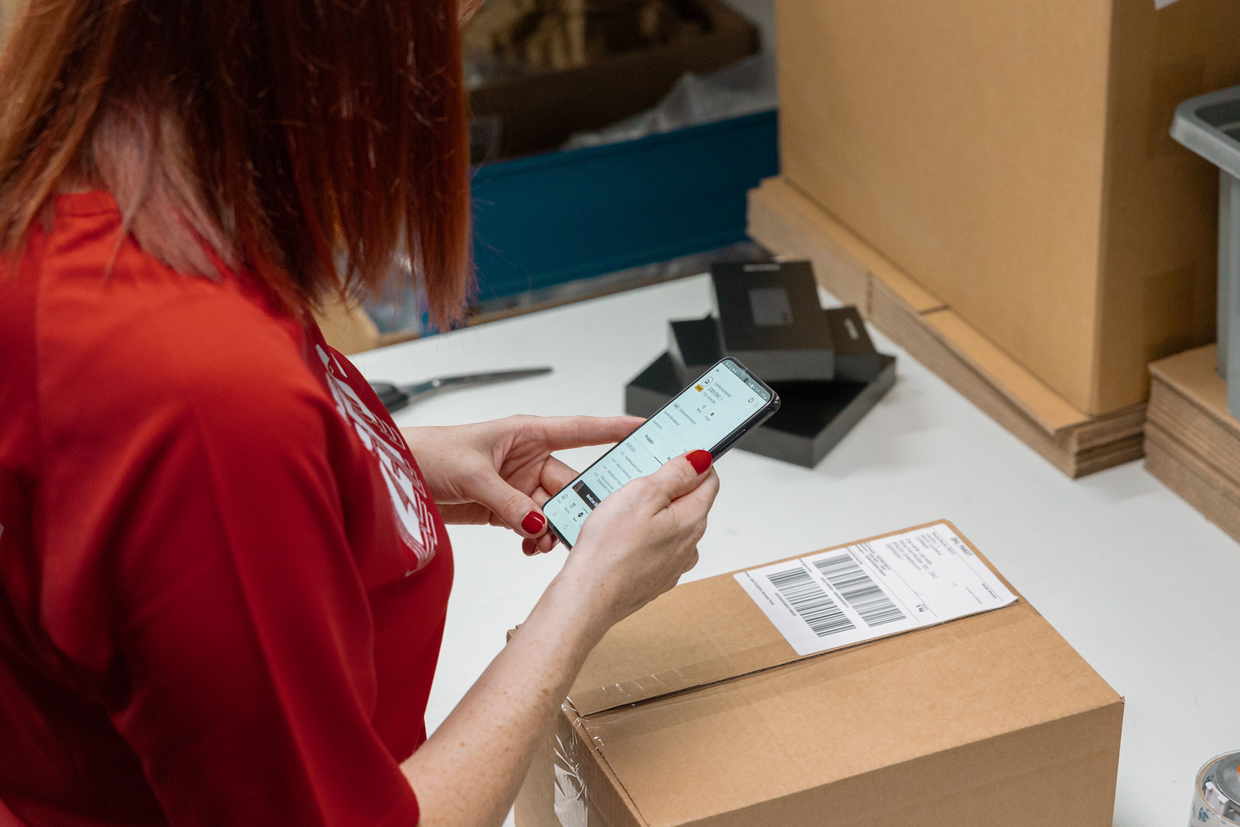 A MediaMarkt employee viewing order information on a smartphone while standing in front of a cardboard box.
