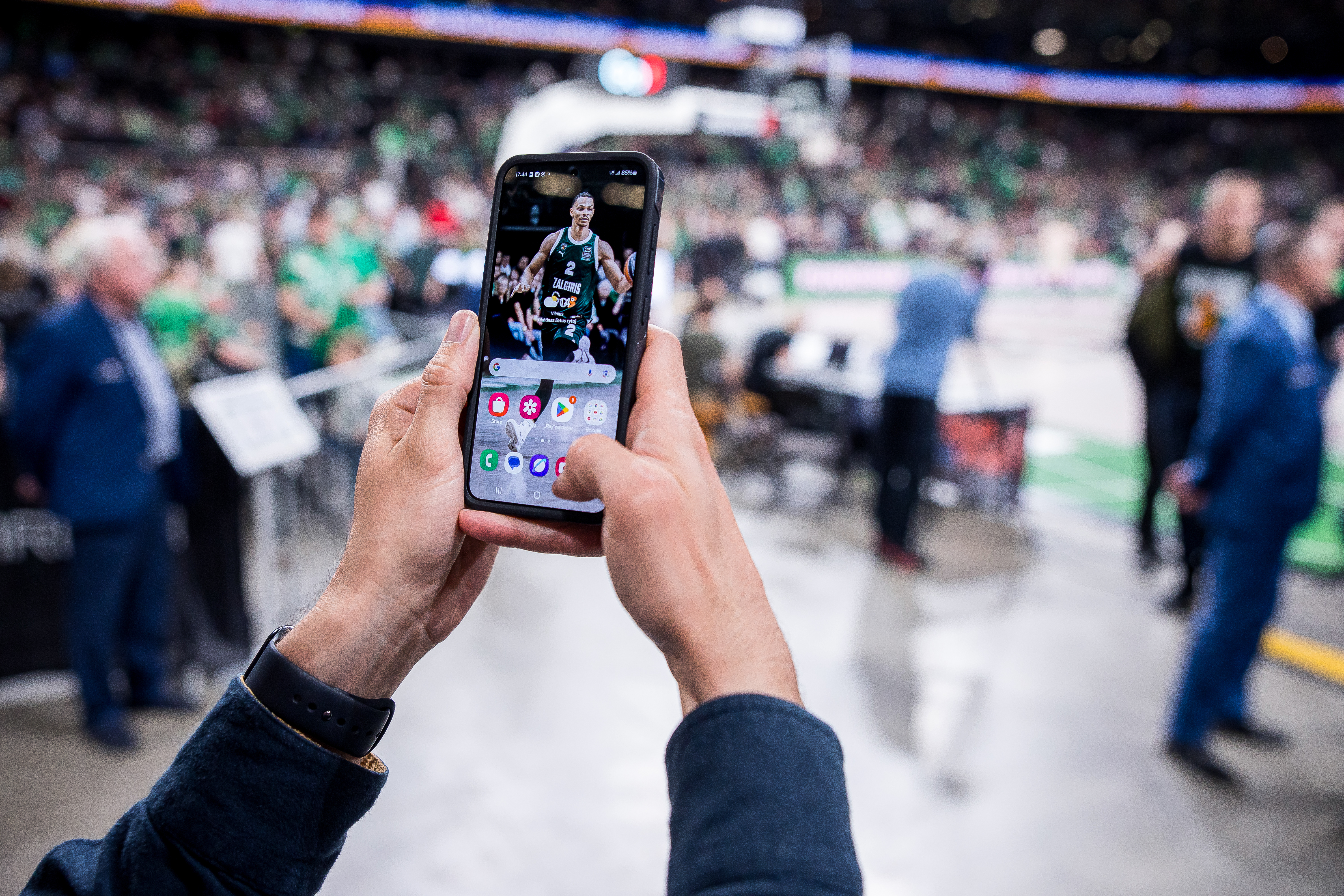 A person holding a Samsung smartphone displaying a Žalgiris basketball player as the wallpaper at a live basketball game.