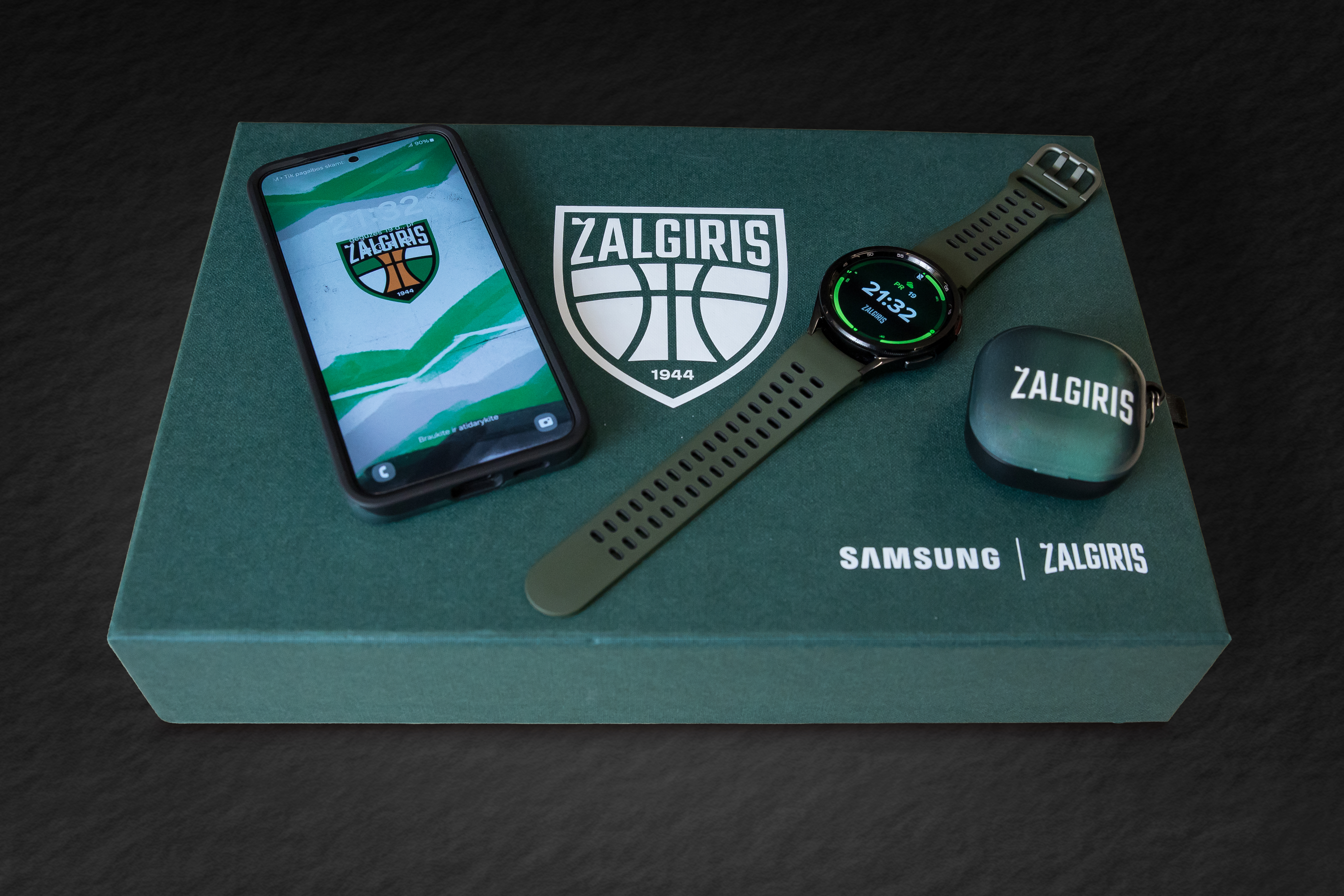 A Samsung and Žalgiris co-branded product display featuring a Samsung Galaxy smartphone with a Žalgiris wallpaper, a green smartwatch, and matching earbuds case arranged on a branded box.